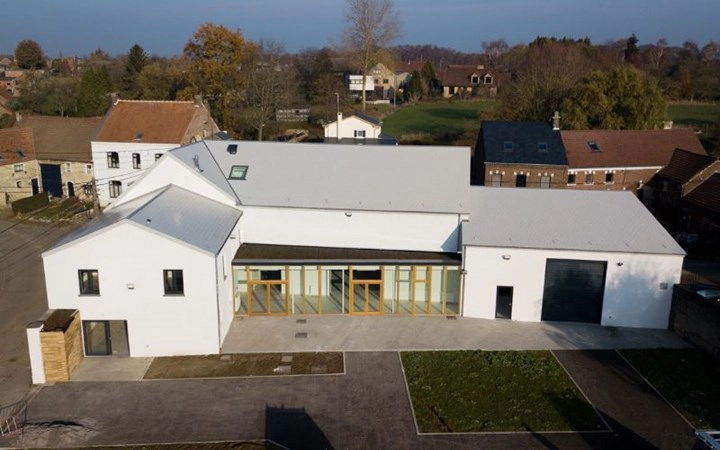 Complexe de bâtiments modernes avec des murs blancs et un toit gris. La structure comprend plusieurs sections connectées, dont un bâtiment principal avec de grandes fenêtres et portes, et une section adjacente avec une grande porte de garage. Les environs incluent des chemins pavés, des espaces verts, et d'autres bâtiments en arrière-plan.