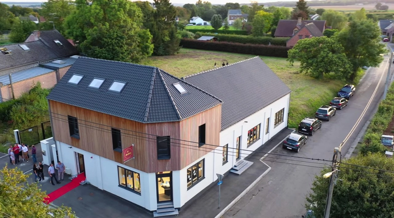 L'image montre un bâtiment moderne à deux étages avec une combinaison de murs extérieurs en bois et blancs. Le bâtiment a un toit incliné avec des puits de lumière et de grandes fenêtres sur les deux étages. Un tapis rouge mène à l'entrée, où plusieurs personnes sont rassemblées. Le bâtiment est situé sur un terrain d'angle avec de la verdure environnante et d'autres maisons en arrière-plan. Plusieurs voitures sont garées le long de la rue à côté du bâtiment.