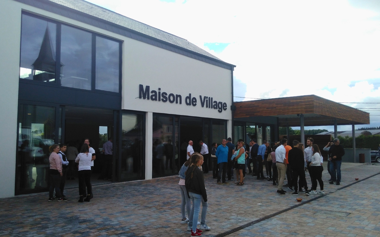 L'image montre un groupe de personnes rassemblées à l'extérieur d'un bâtiment avec le panneau "Maison de Village" dessus. Le bâtiment a de grandes fenêtres et un design moderne, avec un mélange de murs blancs et d'éléments en bois. Le sol est pavé de dalles en pierre, et le ciel semble être partiellement nuageux.