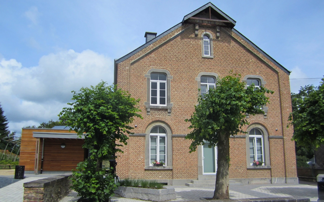 L'image montre une maison en briques à deux étages avec des fenêtres en arc et un toit à pignon. Il y a deux arbres devant la maison, un de chaque côté du chemin d'entrée. La maison a une petite extension en bois sur le côté gauche. Le ciel est partiellement nuageux.