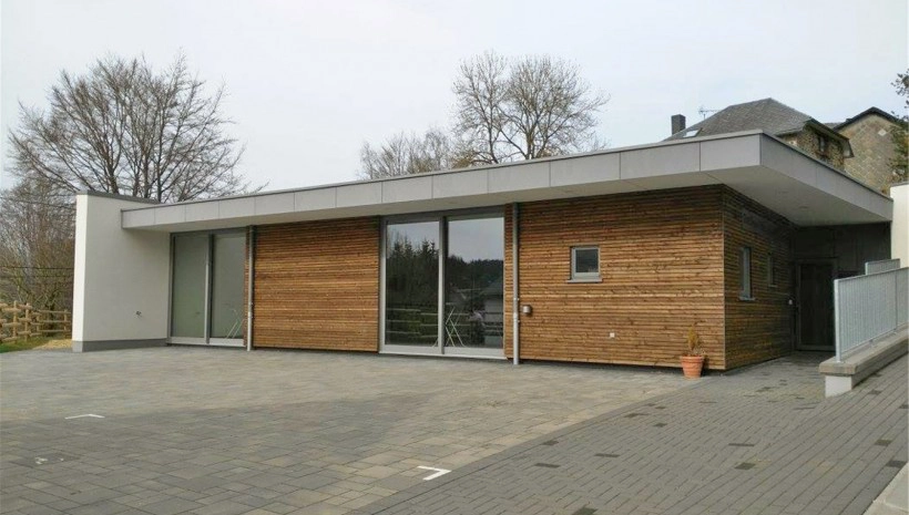 L'image montre un bâtiment moderne d'un seul étage avec un toit plat. Les murs extérieurs sont partiellement recouverts de planches de bois horizontales et de grandes fenêtres/portes vitrées. Il y a une zone pavée devant le bâtiment, et des arbres sont visibles en arrière-plan.