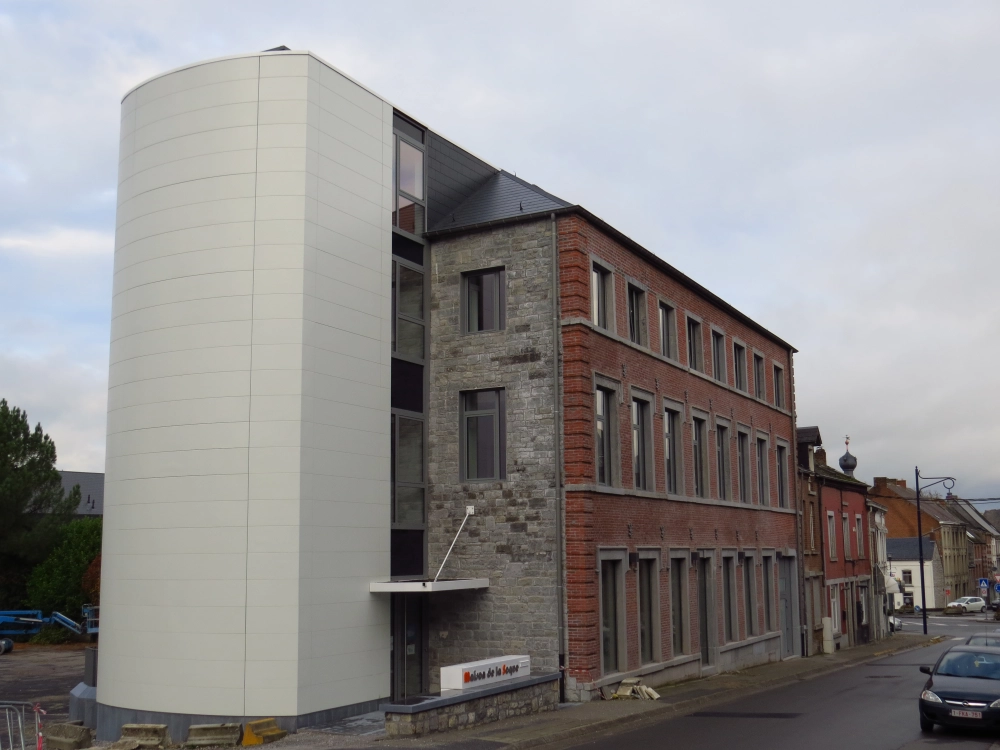 L'image montre un bâtiment à plusieurs étages avec une extension moderne sur le côté gauche. La structure originale est en briques et en pierre, tandis que l'extension a une façade blanche et lisse. Il y a un panneau près de l'entrée qui indique "Maison de la Laïcité". Le bâtiment est situé le long d'une rue avec d'autres bâtiments visibles en arrière-plan.