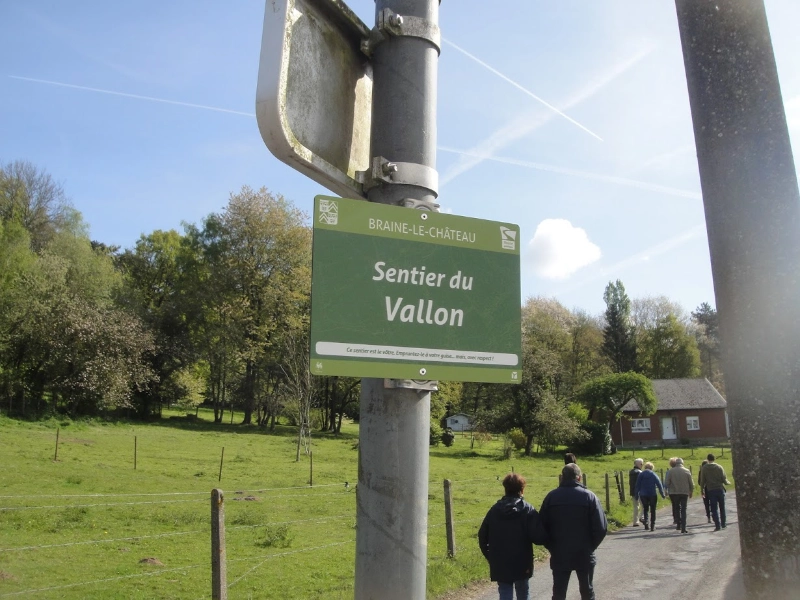 L'image montre un groupe de personnes marchant sur un sentier dans une zone rurale. Il y a un panneau vert attaché à un poteau qui indique : BRAINE-LE-CHÂTEAU Sentier du Vallon Ce sentier est relié à l'itinéraire de la promenade des crêtes (n° 38).