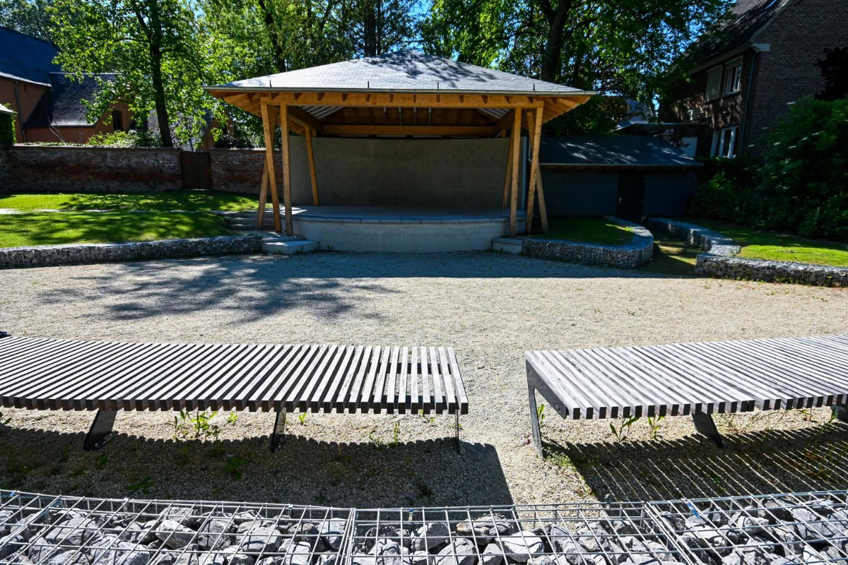 Un amphithéâtre extérieur avec une scène en bois et un toit soutenu par des poutres en bois. Il y a deux longs bancs à lattes au premier plan, et la zone autour de la scène est recouverte de gravier. Des arbres et des bâtiments sont visibles en arrière-plan.