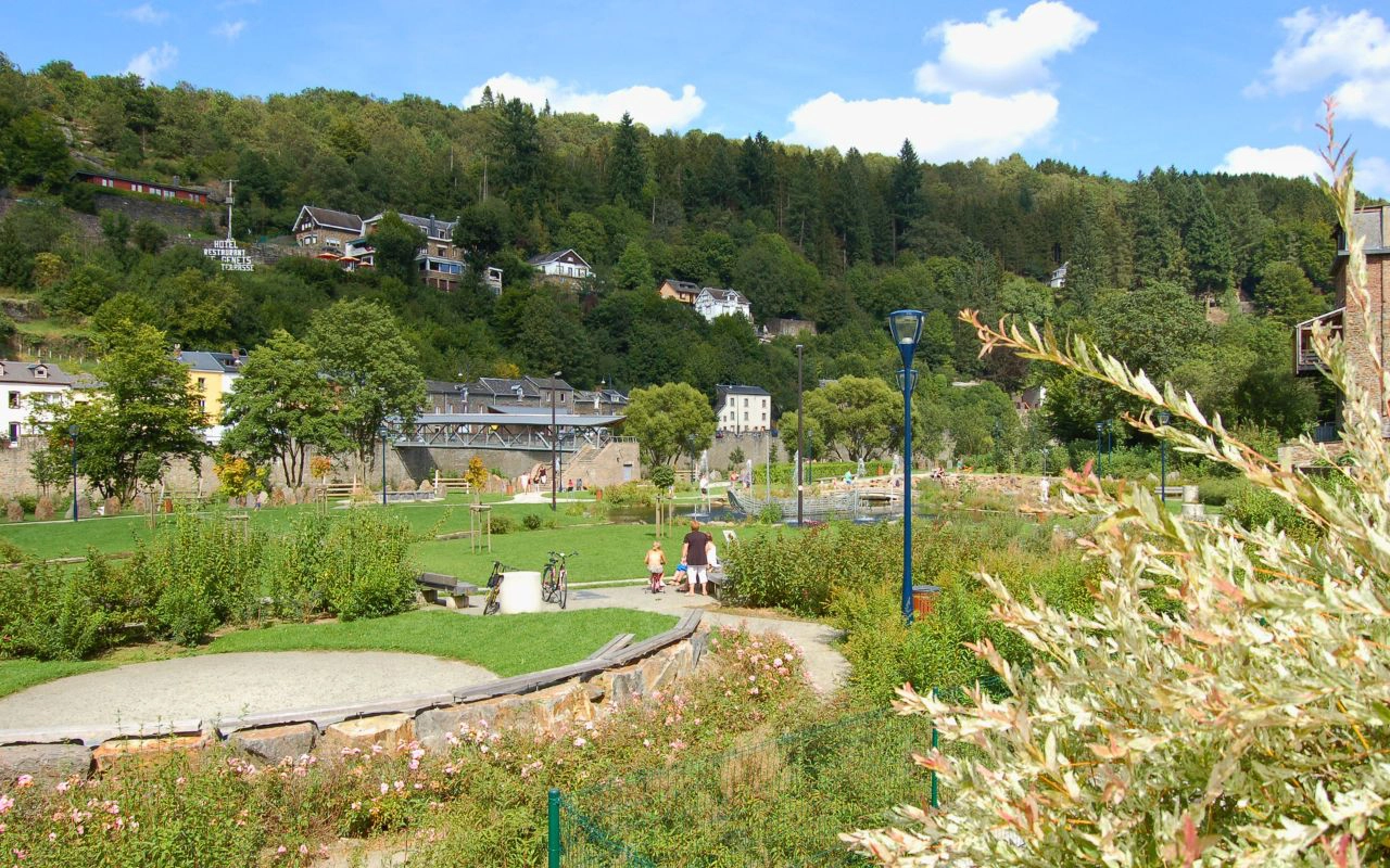 Un parc avec des pelouses vertes, des chemins et diverses plantes. Des personnes marchent le long des chemins. En arrière-plan, il y a des maisons sur une colline couverte d'arbres. Le ciel est bleu avec quelques nuages.