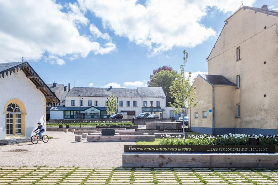 Une place extérieure avec des bancs, des arbres et des bâtiments en arrière-plan. Une personne fait du vélo sur le côté gauche de l'image. Il y a une citation écrite sur un banc en français : "Des souvenirs autour des savoirs... Des rencontres sur (illisible)"