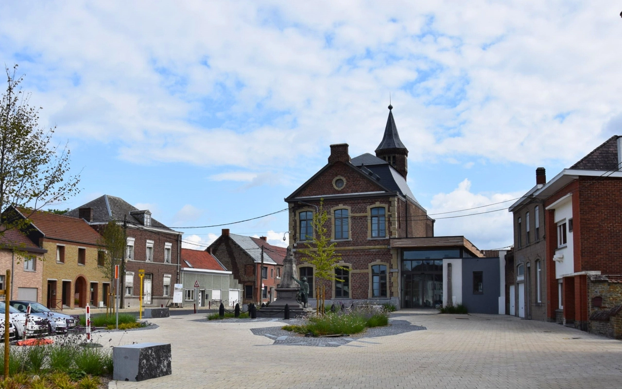 Une petite place de village entourée de plusieurs bâtiments. Le bâtiment central semble être une structure ancienne avec une tour et des fenêtres en arc. Il y a d'autres bâtiments plus petits autour, y compris des maisons. La place elle-même est pavée de dalles en pierre et comporte quelques espaces verts, y compris de petits arbres et des plantes dans des zones désignées.