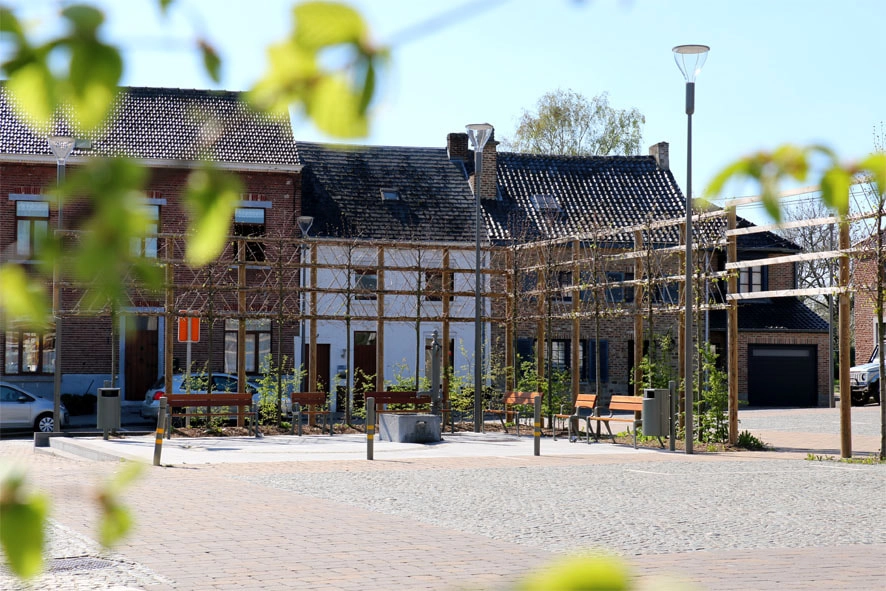 Une petite place de village tranquille avec un pavage en pierre. Il y a plusieurs bancs et de jeunes arbres plantés dans la zone. En arrière-plan, il y a des bâtiments en brique avec des toits en tuiles, dont certains ont des cheminées. Un lampadaire est également visible sur le côté droit de l'image. Au premier plan, des feuilles vertes floues suggèrent que la photo a été prise derrière un arbre ou un buisson.