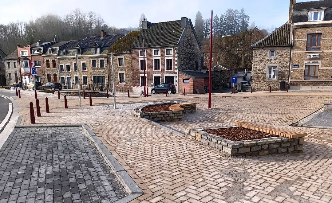 Une place pavée dans ce qui semble être un petit village. La zone est entourée de bâtiments avec des façades en pierre et en brique. Il y a plusieurs poteaux et bornes rouges, ainsi que des jardinières surélevées avec des bancs en bois autour. Le sol est recouvert de pavés autobloquants, et il y a des places de parking marquées sur le côté gauche de l'image.