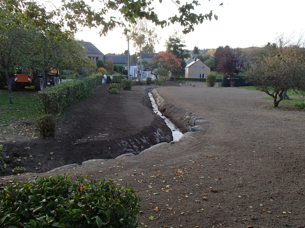 Zone de jardin ou de parc avec un petit ruisseau traversant le milieu. Le sol semble fraîchement labouré ou préparé, probablement pour la plantation. Il y a des arbres et des buissons de chaque côté du ruisseau, et quelques personnes sont visibles en arrière-plan près de bâtiments. Le ciel est dégagé, indiquant qu'il pourrait être en fin d'après-midi ou en début de soirée.