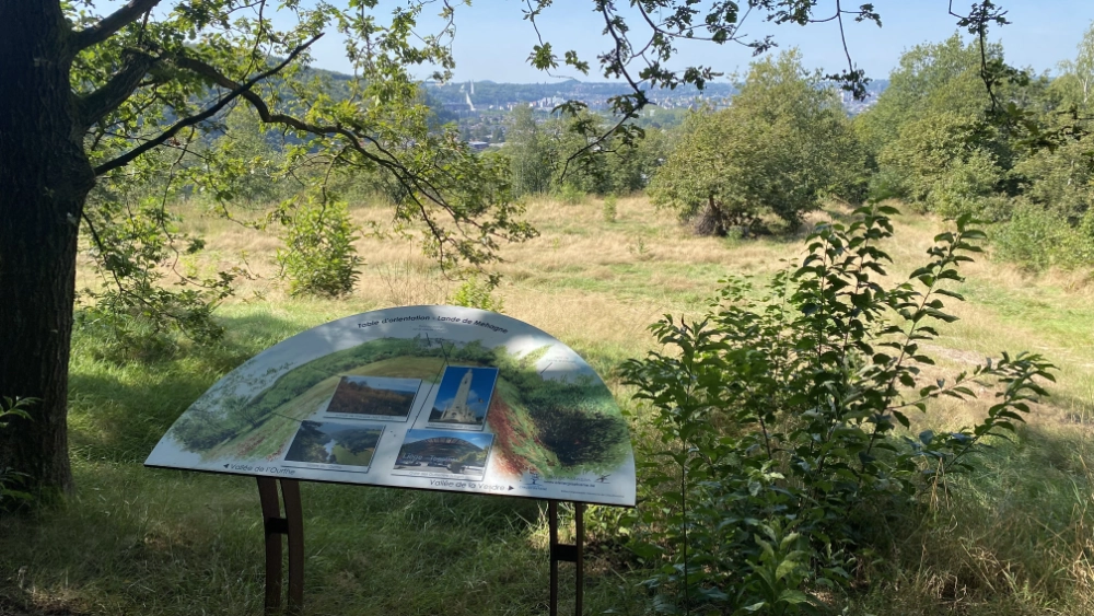 Panneau d'information extérieur situé dans un champ herbeux entouré d'arbres. Le panneau contient plusieurs images et du texte, probablement fournissant des informations sur la région environnante. En arrière-plan, on peut voir davantage d'arbres et un village au loin.