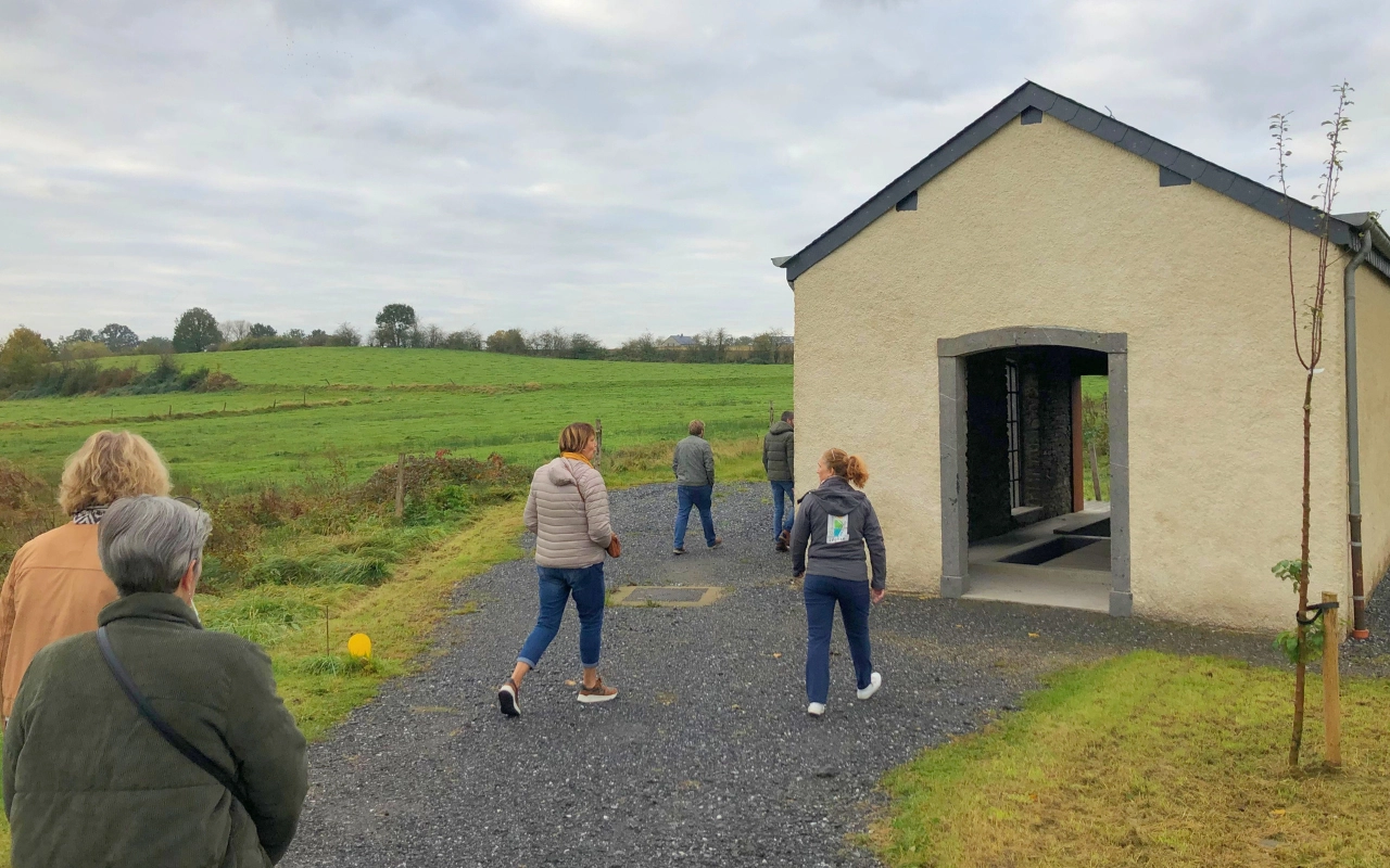 Un groupe de personnes marchant vers un petit bâtiment avec une entrée ouverte. Le bâtiment est situé dans une zone rurale avec des champs verts et des arbres en arrière-plan. Le ciel est nuageux, et le sol est recouvert de gravier.