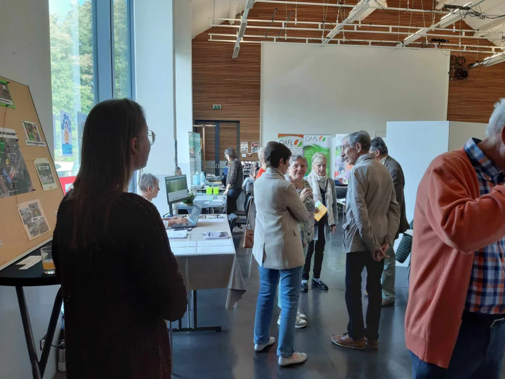 : Un groupe de personnes debout et conversant dans ce qui semble être un espace d'exposition ou un événement intérieur. Il y a des tables avec divers objets et présentoirs, y compris un panneau avec des photos sur le côté gauche. La pièce a de grandes fenêtres laissant entrer la lumière naturelle, et plusieurs affiches et documents d'information sont visibles en arrière-plan.