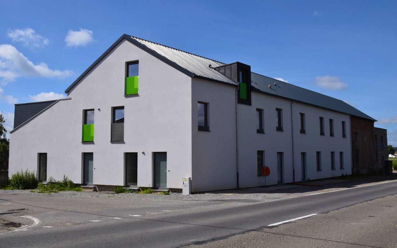 L'image montre un bâtiment moderne à deux étages avec une façade blanche et des accents verts autour de certaines fenêtres. Le bâtiment a un toit en métal incliné et plusieurs fenêtres sur les deux étages. Il est situé à côté d'une route pavée, et il n'y a pas de véhicules ou de personnes visibles dans l'image. Le ciel est dégagé avec quelques nuages épars.