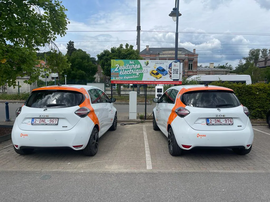 L'image montre deux voitures blanches avec des accents orange garées côte à côte dans des places de parking désignées. Il y a un panneau en arrière-plan qui indique "Les 2 voitures électriques". Le panneau comprend également du texte supplémentaire et des images de voitures.