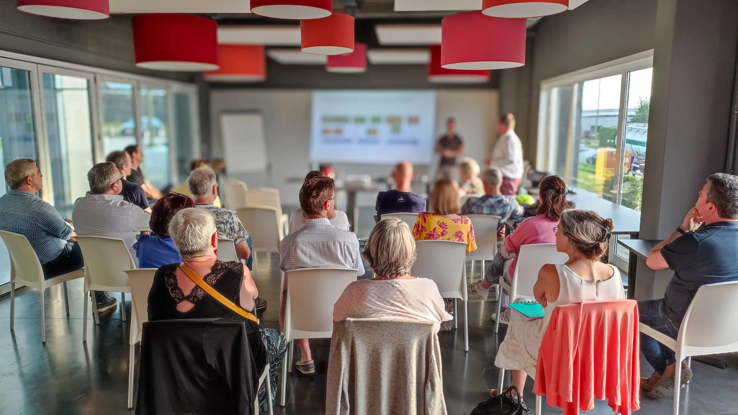 Dans une salle, au premier plan, des personnes assises que l'on voit de dos et qui regardent sur un grand écran la présentation de l'avant-projet du PCDR de leur commune.