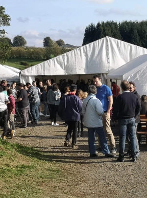 Un groupe de personnes se rassemble autour et à l'intérieur d'un chapiteau blanc installé sur une pelouse. Certaines personnes discutent debout à l'extérieur, tandis que d'autres sont assises à des tables à l'intérieur de la tente. En arrière-plan, on aperçoit des arbres, des collines et un ciel partiellement nuageux.