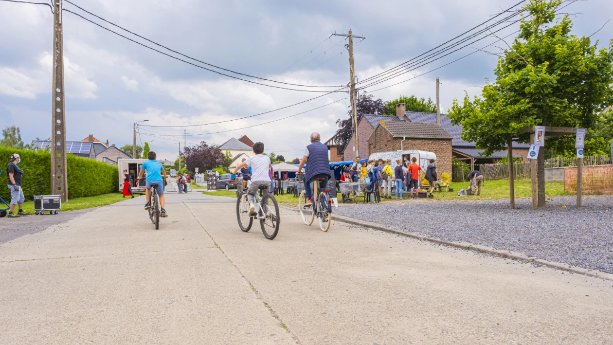 Scène de vie dans un village wallon : plusieurs personnes circulent à vélo sur une route en béton, tandis qu’un groupe se rassemble autour d’un stand installé devant des maisons en briques.