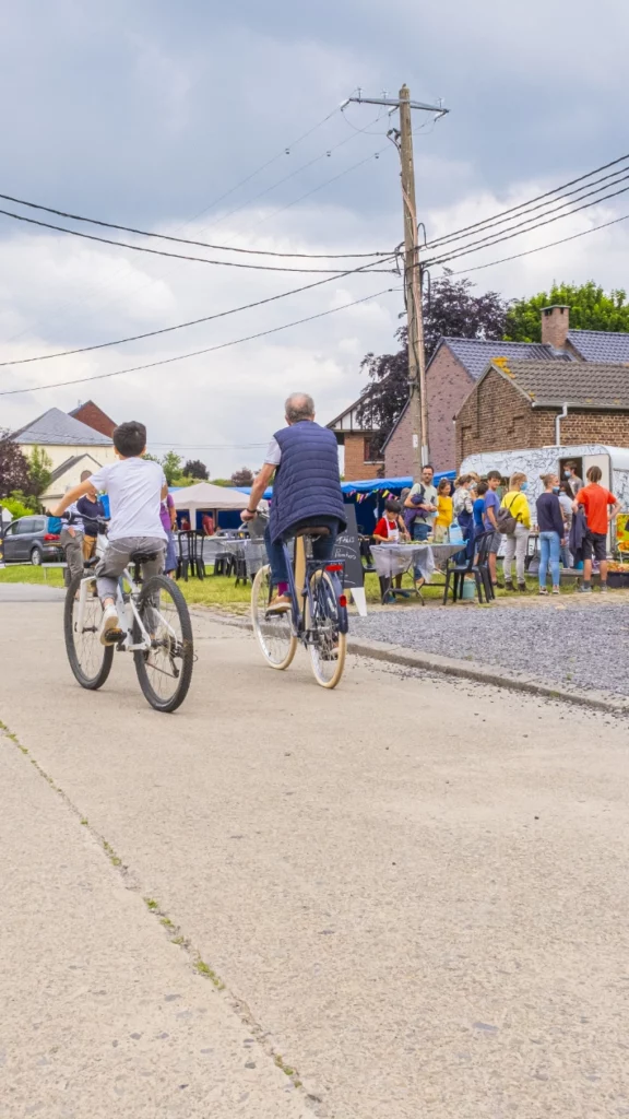 Scène de vie dans un village wallon : plusieurs personnes circulent à vélo sur une route en béton, tandis qu’un groupe se rassemble autour d’un stand installé devant des maisons en briques.