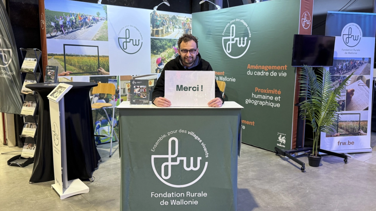 Sur le stand de la Fondation Rurale de Wallonie au Salon des Mandataires, une personne est assise derrière un comptoir aux couleurs de la FRW et tient une affiche « Merci ! ». À l’arrière-plan, des panneaux de communication présentent le logo de la FRW, des visuels et des slogans. 