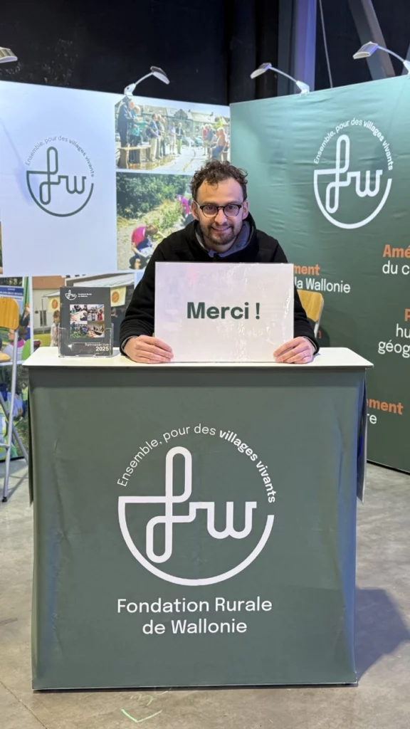 Sur le stand de la Fondation Rurale de Wallonie au Salon des Mandataires, une personne est assise derrière un comptoir aux couleurs de la FRW et tient une affiche « Merci ! ». À l’arrière-plan, des panneaux de communication présentent le logo de la FRW, des visuels et des slogans.