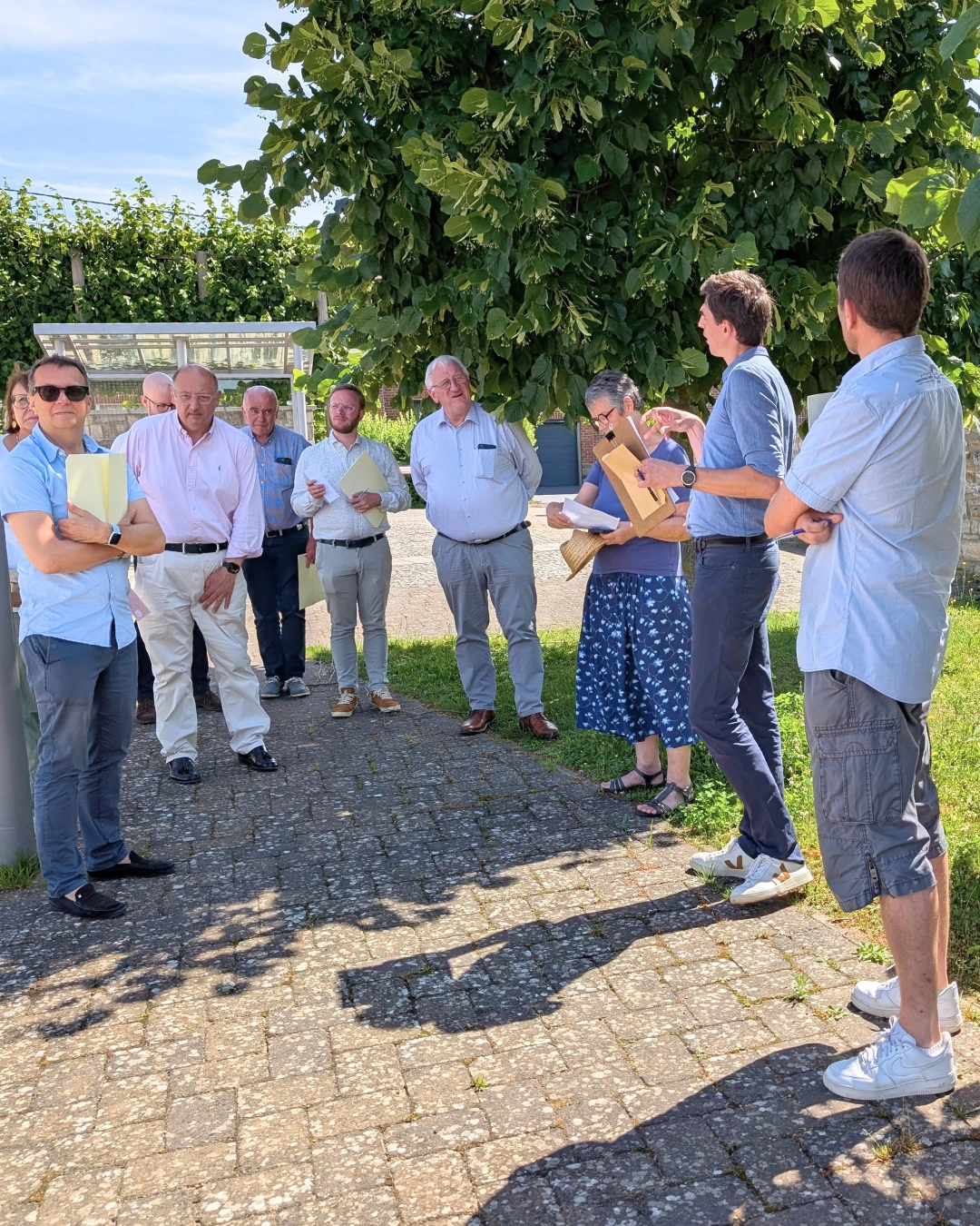 Groupe de participants réunis en extérieur lors d'une visite de terrain, échangeant autour d'un projet local sous un grand arbre par une journée ensoleillée.